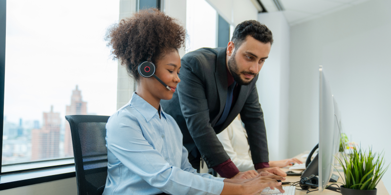 Mulher atendente com headset trabalhando em frente ao computador enquanto recebe orientação de colega.